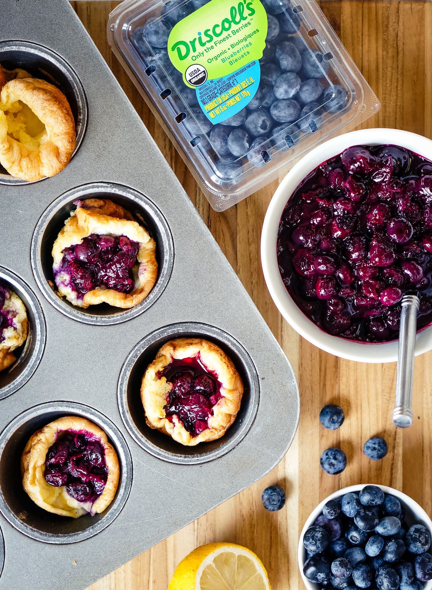 Dutch babies still in the muffin pan next to blueberry filling, a Driscoll's organic blueberry clamshell, and a lemon on top of a wood countertop.