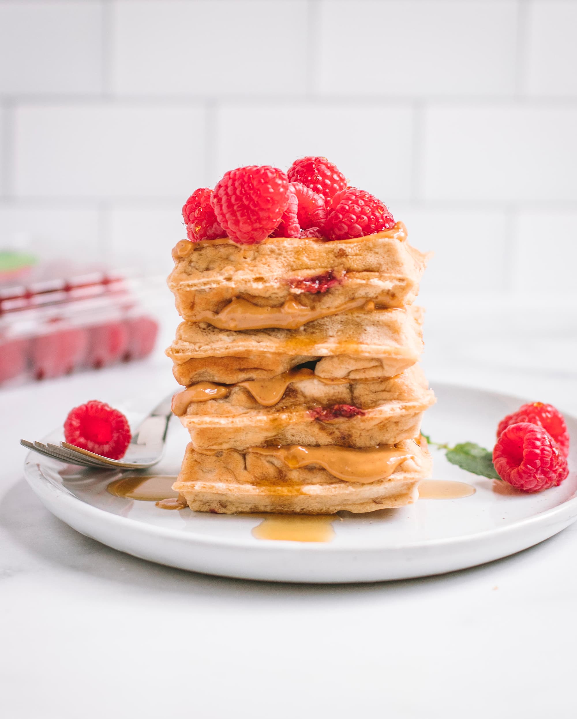 Peanut butter waffles topped with raspberries. A Driscoll's raspberry clamshell is sitting on the counter behind the plate.