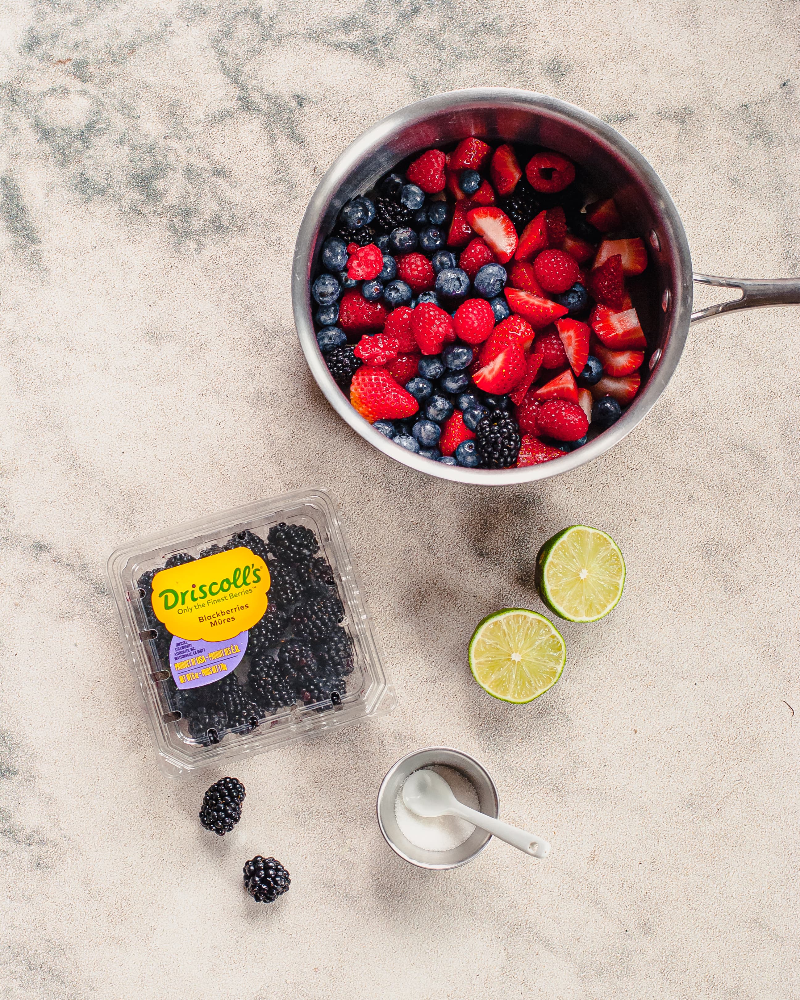 Top view of ingredients needed to make the panna cotta berry compote. Raspberries, strawberries, blackberries, and blueberries in a pot with a lime and sugar next to it