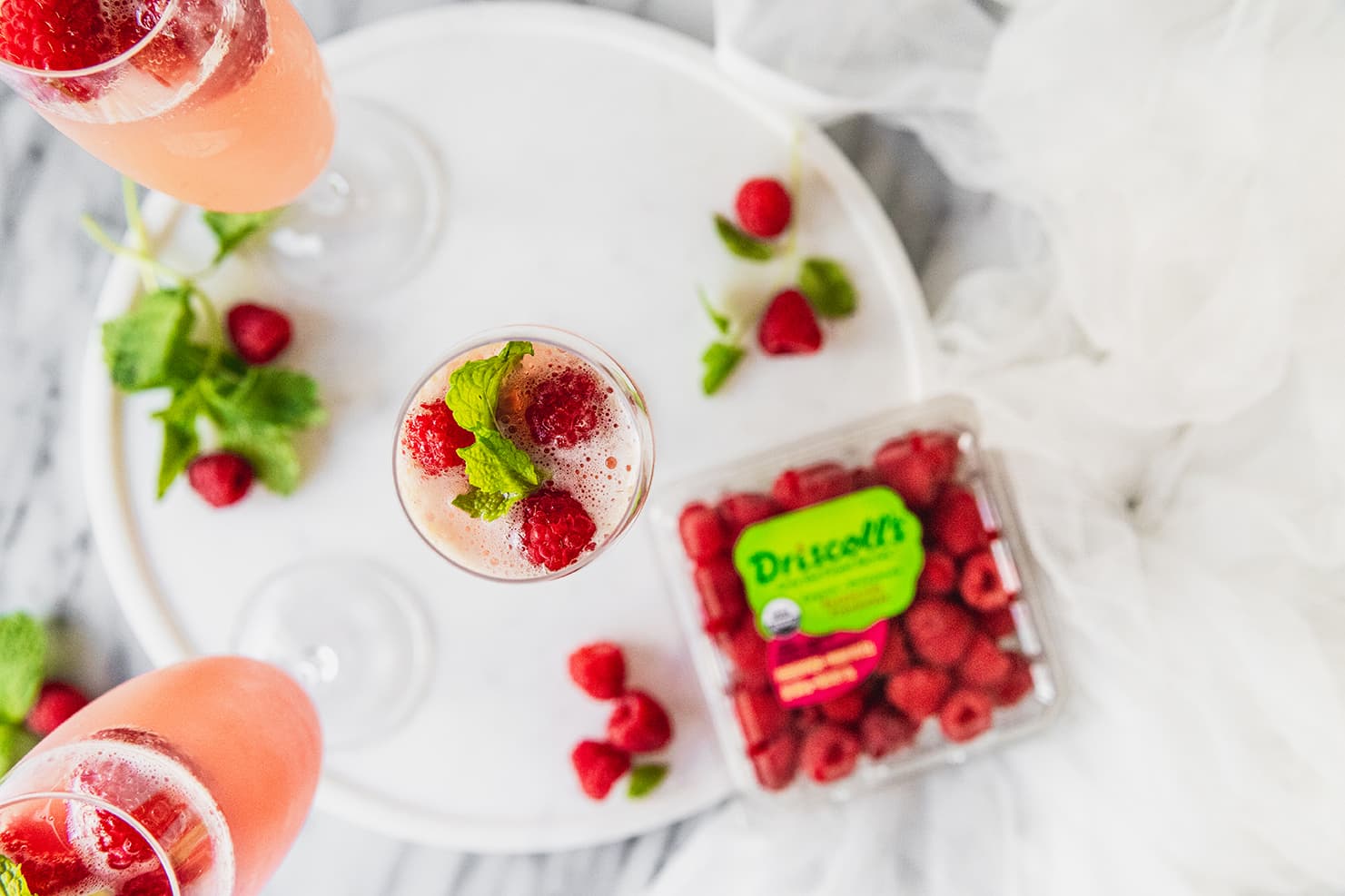Top-down view of raspberry mimosas topped with fresh raspberries and garnish. A Driscoll's clamshell of organic raspberries sit at the base of one of the champagne glasses. The raspberry mimosas are sitting on a white plate on top of a granite countertop.