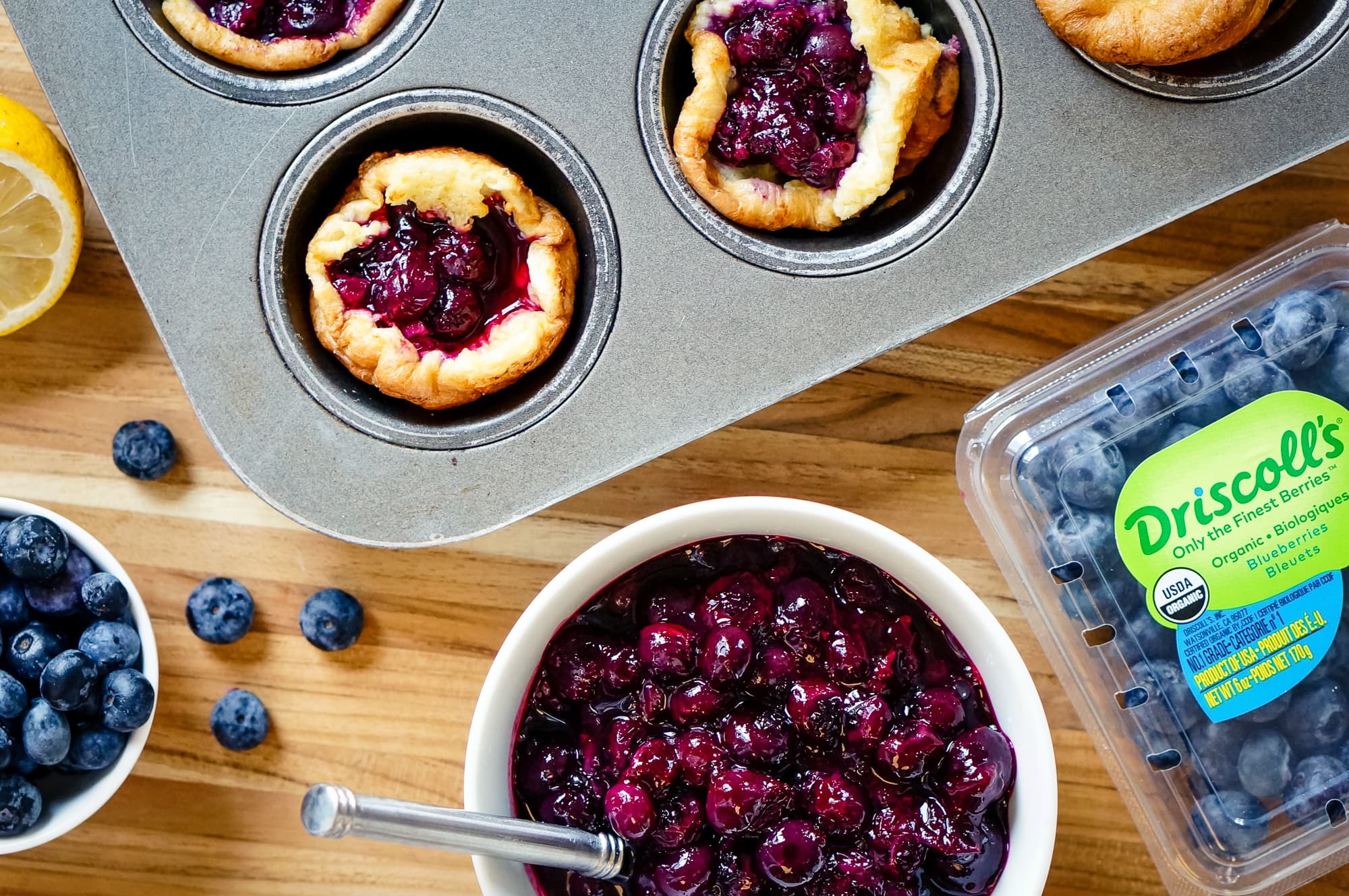 Dutch babies still in the muffin pan next to blueberry filling, a Driscoll's organic blueberry clamshell, and a lemon on top of a wood countertop.
