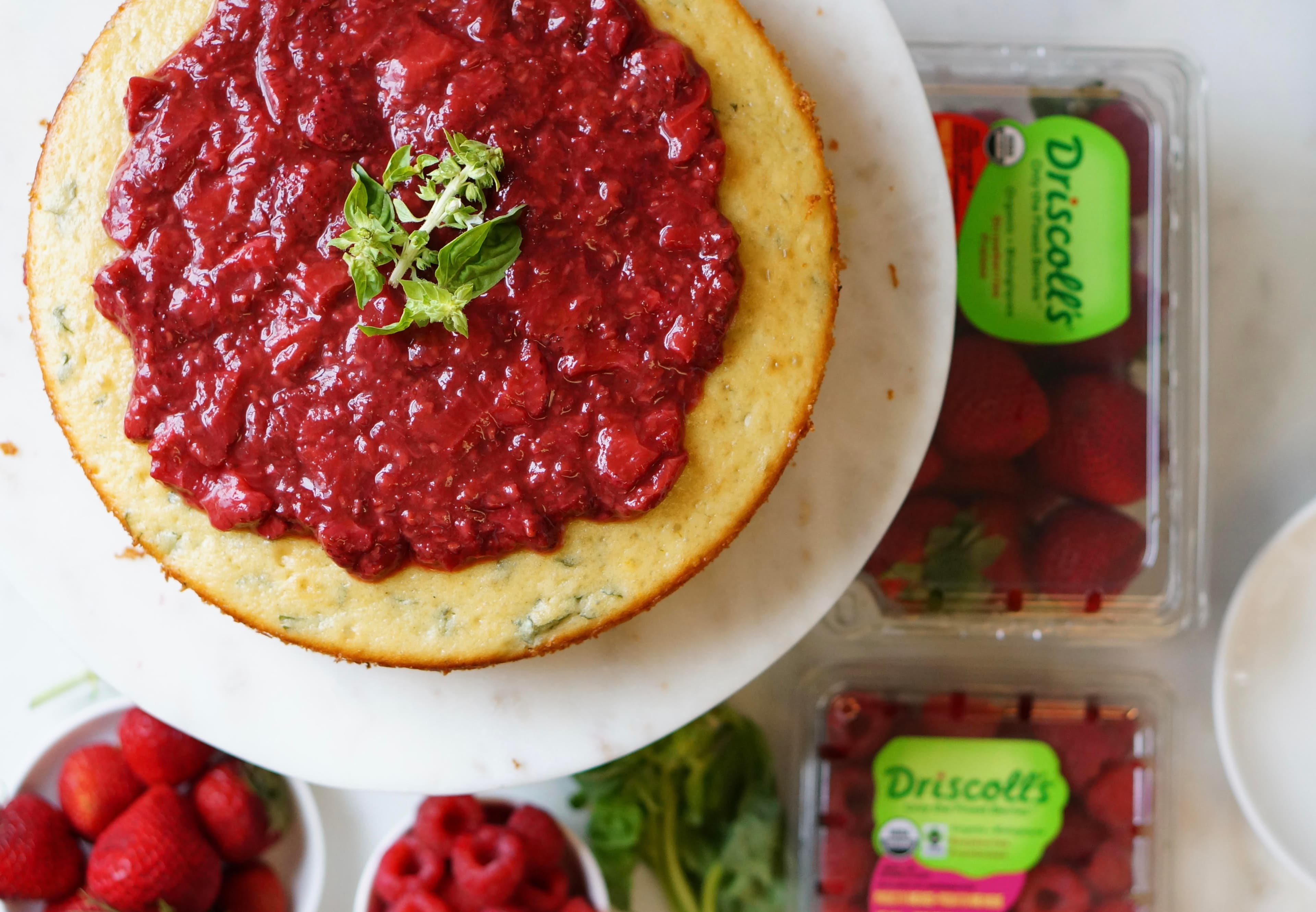 Top view of a strawberry raspberry basil cake top with a strawberry raspberry compote and basil garnish. Plate surrounded by bowls of fresh raspberries and strawberries.