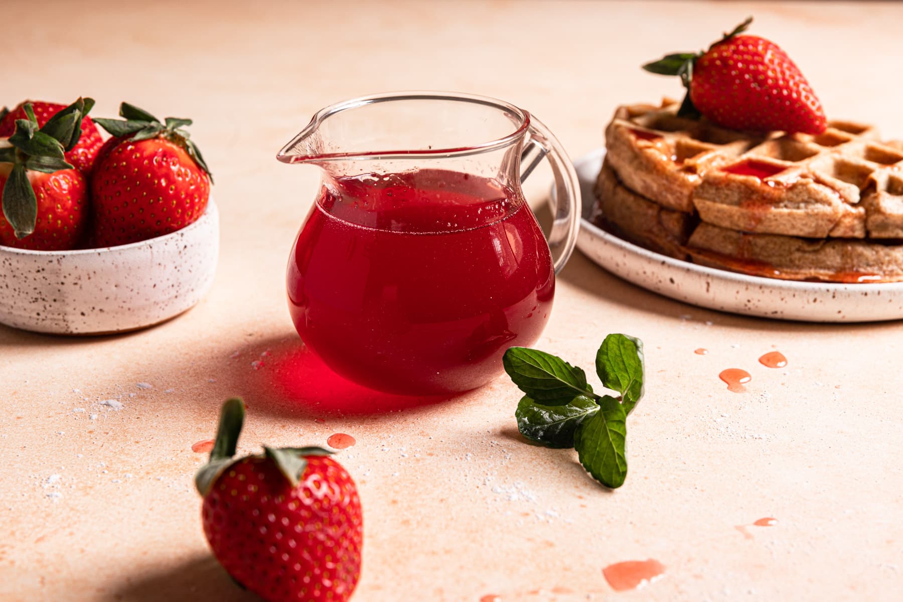 A pitcher of strawberry basil syrup sitting next to a plate of waffles.